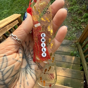 May include: A clear resin bookmark filled with gold glitter, featuring a red tassel and various black and gold icons including glasses, footprints, a hat, and a lion crest. The name EMILY is spelled out vertically in white beads with black letters.
