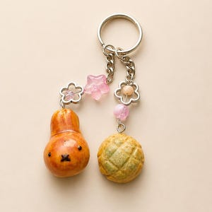 May include: A silver-toned metal keychain featuring two dangling charms: a glossy, orange-brown pear-shaped character with a simple face and a textured, golden-brown round bread roll. The charms are accented with pink star and flower beads.