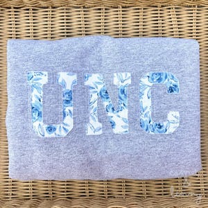 Puede incluir: Una sudadera gris jaspeado con las letras UNC en fuente de bloque. Las letras están rellenas con un patrón de tela blanca que muestra ilustraciones florales azules de rosas y hojas, creando un contraste marcado con la tela gris.