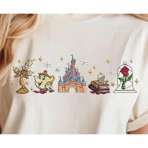 May include: A cream-colored t-shirt featuring an embroidered design of a golden candelabra, a smiling teapot, a pink and blue castle, a stack of books with a candle, and a red rose inside a glass dome, accented with small stars and white flowers.