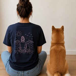 Peut inclure: Un t-shirt bleu marine avec un motif en lignes rose clair au dos. Le dessin représente une personne coiffée d'un chignon assise à côté d'un chien, face à un paysage composé de montagnes, d'un soleil et d'oiseaux volant au-dessus de l'eau.