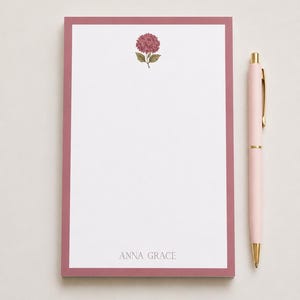 May include: A rectangular notepad with a white center and a dusty rose border. The top features a small illustration of a dark pink hydrangea flower with green leaves. The name ANNA GRACE is printed in gray at the bottom. A light pink pen with gold accents.