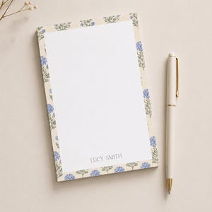 May include: A rectangular notepad with a white center and a border featuring a repeating pattern of blue hydrangea flowers on a cream background. The name LUCY SMITH is printed in gray at the bottom center of the white area, accompanied by a cream pen.