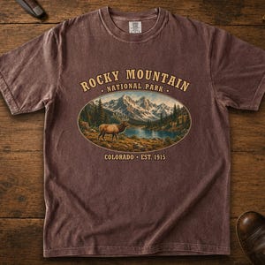 Puede incluir: Una camiseta de manga corta de color marrón desteñido con un gráfico ovalado central de un paisaje montañoso con un alce, un lago y pinos. El texto dice ROCKY MOUNTAIN NATIONAL PARK y COLORADO EST. 1915 en una tipografía de color beige.