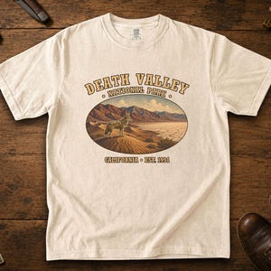 Puede incluir: Una camiseta de manga corta color crema con un gráfico de un coyote de pie sobre dunas de arena en el Parque Nacional del Valle de la Muerte. El texto dice DEATH VALLEY NATIONAL PARK y CALIFORNIA EST. 1994 en una fuente marrón arqueada.