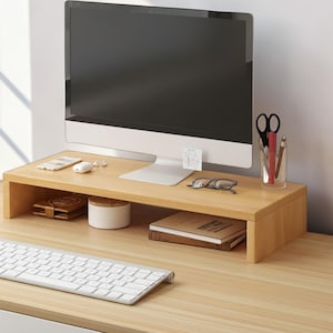 Peut inclure: Un support d'écran en bois clair avec un plateau rectangulaire et deux supports latéraux. Le support accueille un écran d'ordinateur, un pot à crayons transparent avec des ciseaux et des stylos, des lunettes, une petite note blanche et un smartphone.