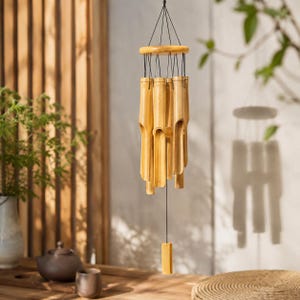 Op de afbeelding: Een hangend windgong gemaakt van meerdere lichtbruine bamboebuizen van verschillende lengtes. De buizen hangen aan een ronde houten ring met zwarte koorden, met een enkel rechthoekig bamboe stuk dat in het midden naar beneden hangt.
