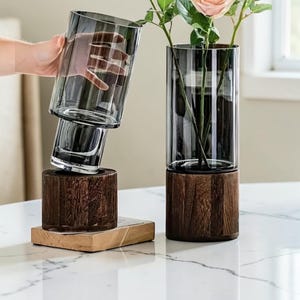 Könnte beinhalten: Zwei zylindrische Glasvasen mit dunkelgrauer Tönung, die jeweils in einem runden, dunkelbraunen Holzsockel stecken. Eine Vase wird schräg über ihrem Sockel gehalten, während die andere aufrecht steht und einen künstlichen rosafarbenen Rosenstiel enthält.