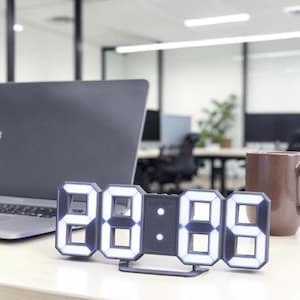 Puede incluir: Un reloj digital rectangular con marco gris oscuro y segmentos LED de color blanco brillante que muestran la hora. El reloj cuenta con un panel central con dos pequeñas luces circulares y el texto "MOM Store" impreso en letras gris oscuro.