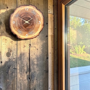 May include: A round wall clock crafted from a natural wood slice featuring visible tree rings and bark edges. The clock face displays white numerals at the 12, 3, 6, and 9 o'clock positions, complemented by thin, gold-coloured metal hour and minute hands.
