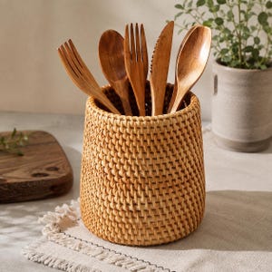 Può includere: Un portautensili cilindrico in rattan intrecciato contenente un set di posate in legno, tra cui due forchette, due cucchiai e un coltello seghettato. Il contenitore in vimini marrone chiaro presenta una trama fitta, uniforme e solida.