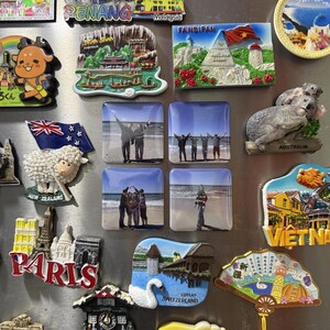 Puede incluir: Una colección de varios imanes de nevera de recuerdo con diversos diseños, incluyendo una oveja de Nueva Zelanda, un paisaje de montaña Fansipan, un koala, un conjunto de monumentos de París y cuatro imanes cuadrados con fotos personales.
