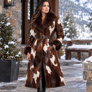 May include: A long, calf-length coat featuring a brown and white cow-print pattern. The garment includes a dark brown fur collar and matching fur cuffs at the wrists. A dark brown belt with a rectangular buckle cinches the waist of the coat.