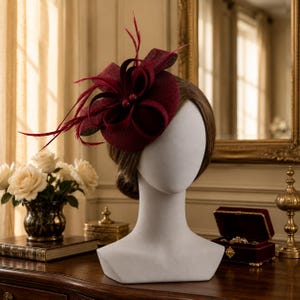 Könnte beinhalten: Ein tiefburgunderroter Fascinator mit einer strukturierten kreisförmigen Basis, kunstvollen Stoffschleifen und zarten, farblich abgestimmten Federn. Das Kopfstück wird auf einem neutralen Mannequinkopf präsentiert und zeigt seine Textur.