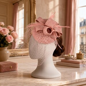 Op de afbeelding: Een oudroze fascinator met een gestructureerde ronde basis, een grote strik en een bijpassende netsluier. Het hoofddeksel is versierd met dunne, fijne veren die vanuit het midden uitsteken en wordt gepresenteerd op een etalagepop.