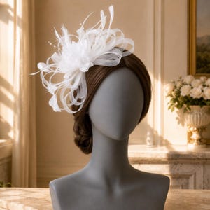 May include: A white fascinator headpiece featuring layered sheer fabric loops, delicate feathers, and small white beads. The accessory is positioned on the side of a grey mannequin head, showcasing a structured design with airy, curved elements.