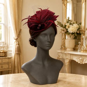 Peut inclure: Un bibi de couleur bordeaux profond orné de boucles en tissu structuré, de plumes délicates et d'un voile en résille fine. L'accessoire est présenté sur un buste de mannequin gris foncé, mettant en valeur son design complexe et superposé.