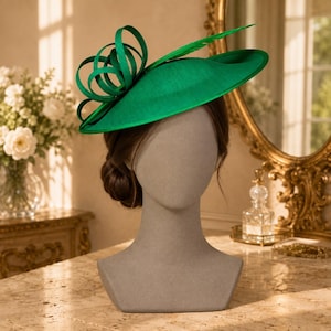 Pode incluir: Um fascinator verde esmeralda vibrante com uma aba larga e plana e um laço escultural elaborado feito de laços de fita verde a condizer. O chapéu é exibido numa cabeça de manequim de tom neutro, destacando o seu design estruturado e textura.