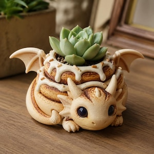 May include: A light beige dragon-shaped planter featuring a coiled body, small wings, and a textured surface resembling a cinnamon roll with white icing. A green succulent plant is potted in the center opening of the dragon's back.