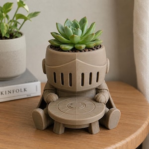 May include: A light brown, 3D-printed planter shaped like a seated knight wearing a helmet with vertical visor slits. The knight holds a small, round table in front of its lap, and a green succulent plant is potted inside the helmet cavity.