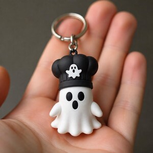May include: A white ghost keychain featuring a black chef hat with a small white ghost icon on the front. The ghost has two black oval eyes and a black oval mouth. The figure is attached to a silver metal split ring for keys.