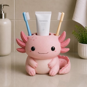 May include: A pink axolotl-shaped toothbrush and toothpaste holder with a smooth finish. The holder features a central slot for a tube of toothpaste labeled TOOTHPASTE WHITENING FRESH MINT and two side slots holding a blue and a yellow toothbrush.