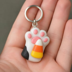 May include: A white cat paw keychain with pink pads and a black spot on the side. A candy corn piece is attached to the center of the paw. The keychain features a silver metal ring at the top for attachment, providing a secure way to hold keys.