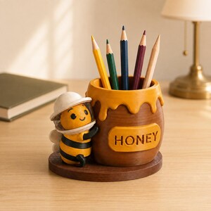 May include: A brown honey pot pencil holder with a yellow bee figure wearing a white beekeeper hat attached to the side. The pot features a dripping honey design at the top and the word HONEY embossed on the front. Five coloured pencils are inside.