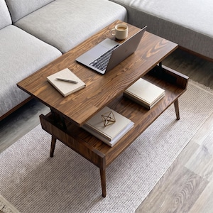 May include: A rectangular wooden coffee table with a lift-top mechanism, featuring a dark brown wood grain finish and four tapered legs. The top surface holds a silver laptop, a white mug, and a notebook with a pen. A lower shelf holds books.