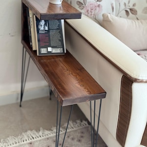 Può includere: Un tavolino stretto in legno marrone scuro con due ripiani orizzontali e sottili gambe a forcina in metallo nero. Il ripiano superiore sostiene una tazza in ceramica bianca e il ripiano inferiore contiene diversi libri disposti in fila.