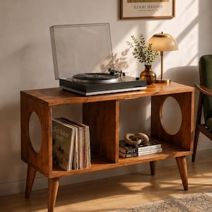 Peut inclure: Une console en bois pour tourne-disque avec quatre pieds fuselés, dotée de deux découpes latérales circulaires et de deux compartiments de rangement ouverts. Le plateau supérieur accueille une platine noire avec un capot transparent.