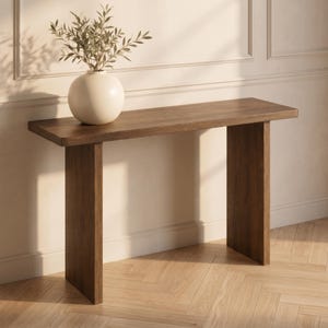May include: A rectangular dark brown wooden console table with a flat top and two vertical slab legs. A round off-white ceramic vase containing a green leafy plant with thin stems sits on the left side of the table surface.