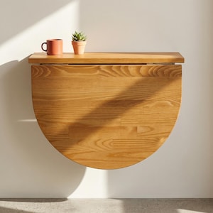 Op de afbeelding: Een aan de muur gemonteerde, halfronde houten klaptafel met een natuurlijke lichtbruine houtnerfafwerking. Op het platte bovenoppervlak van de tafel staan een koraalkleurige keramische mok en een kleine groene vetplant in een terracotta pot.
