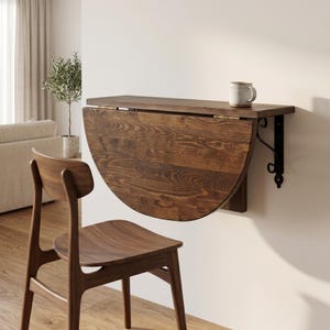 May include: A wall-mounted, semi-circular wooden drop-leaf table with a dark brown wood grain finish, supported by black metal brackets. A light-colored ceramic mug sits on the flat top surface, and a wooden chair is positioned in front.