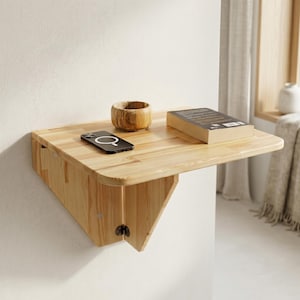Può includere: Una scrivania pieghevole da parete in legno chiaro con superficie liscia, dotata di una piccola ciotola rotonda in legno e uno smartphone nero con design dell'obiettivo circolare, accanto a un libro rettangolare chiuso con copertina scura.