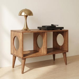May include: A wooden media console with four tapered legs and three open compartments, each featuring a circular cutout on the side panels. A gold-toned mushroom-shaped table lamp and a black record player sit on the top surface of the console.