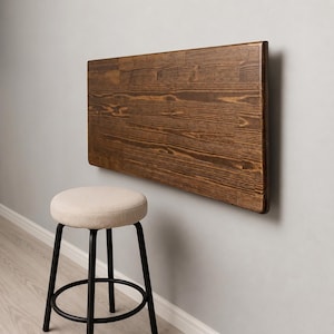 Peut inclure: Une table murale rectangulaire en bois brun foncé avec un grain visible et une finition lisse, placée au-dessus d'un tabouret rond doté d'une assise beige rembourrée et d'un cadre en métal noir à quatre pieds avec un repose-pieds circulaire.
