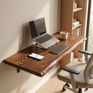 May include: A wall-mounted floating desk featuring a dark brown wood slab with a natural live edge. The desk surface holds a silver laptop on a stand, a wireless keyboard, a smartphone, and a beige ceramic mug. A light-coloured office chair sits nearby.