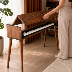 May include: A dark brown wooden piano console featuring a hinged lid that opens to reveal a standard black and white keyboard. The instrument is supported by four tapered wooden legs and has a rectangular frame with a gabled top design.