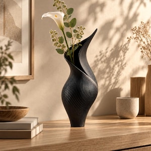May include: A matte black vase with a tall, slender, and curved silhouette featuring a textured, ribbed surface pattern. The vase holds a single white calla lily flower with green stems and small leaves extending from the top opening.