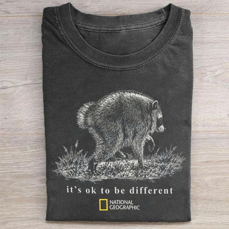 May include: A charcoal gray crew neck t-shirt featuring a white line-art illustration of a raccoon walking through grass. Below the graphic, the text reads "it's ok to be different" followed by the National Geographic logo and brand name.