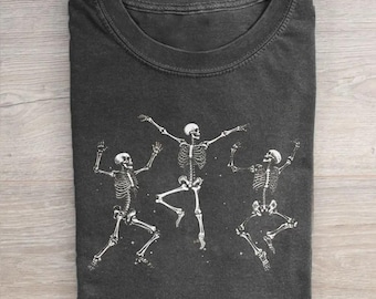 May include: A charcoal gray crew neck t-shirt featuring a white graphic print of three dancing skeletons. The skeletons are depicted in various poses with arms and legs extended, creating a dynamic composition across the front of the garment.