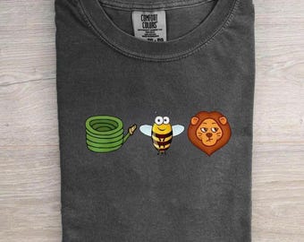 May include: A charcoal gray crew neck t-shirt featuring a graphic print of a green coiled garden hose, a yellow and black striped bee, and a brown lion face. The inner neck label displays the text Comfort Colors with size and care information.