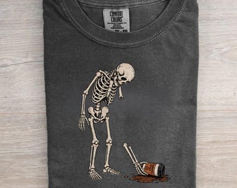 May include: A charcoal gray t-shirt featuring a graphic of a skeleton looking down at a spilled coffee cup on the floor. The skeleton is standing with one arm reaching toward the cup. The text "Well, shit..." is printed in white below the graphic.