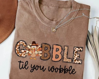 May include: A brown crew neck t-shirt featuring a graphic of a turkey wearing a hat, with the word GOBBLE in large, patterned letters and the text til you wobble written in black script underneath the turkey and the word GOBBLE.
