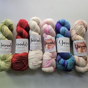 May include: Six skeins of yarn arranged in a row, each with a white paper label reading Yarnable by Hypnotic Yarn. The colors include light green, solid red, cream with speckles, pale pink with speckles, blue and purple gradient, and cream with pink.
