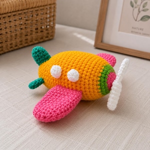 May include: A crocheted toy airplane featuring a bright yellow body, pink wings, and a green tail section. The airplane has two white circular windows on the side, a green accent near the front, and a white propeller attached to the nose.