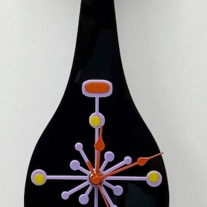 May include: A black cat-shaped wall clock with a glossy finish. The cat has teal eyes and a pink nose. A starburst-shaped clock face in lavender and yellow is centered on the body, featuring orange clock hands and a long, thin black tail.