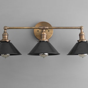 May include: A three-light vanity light fixture with a brass finish and black metal shades. The fixture has a straight bar design with three arms extending from the center.