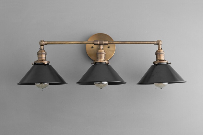 May include: A three-light vanity light fixture with a brass finish and black metal shades. The fixture has a straight bar design with three arms extending from the center.